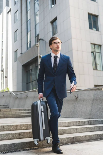 Joven hombre de negocios guapo en traje elegante con equipaje y billetes de avión bajando escaleras en el distrito de negocios - foto de stock