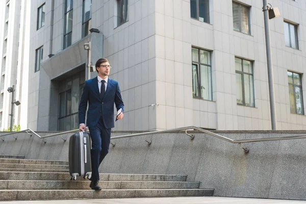 Hombre de negocios en traje elegante con equipaje y billetes de avión que bajan escaleras en el distrito de negocios - foto de stock