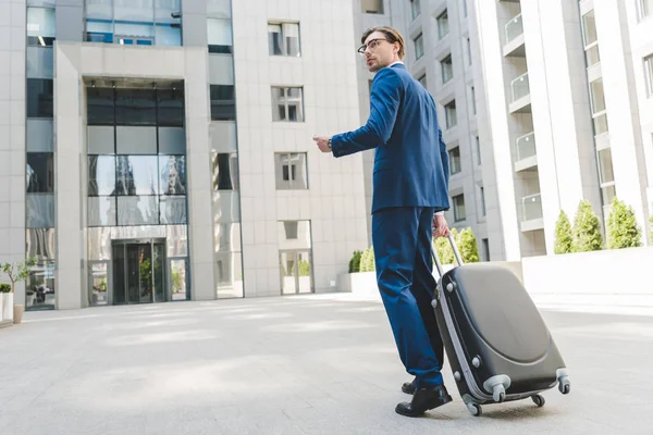 Joven hombre de negocios guapo en traje elegante con equipaje y billetes de avión caminando por el distrito de negocios - foto de stock
