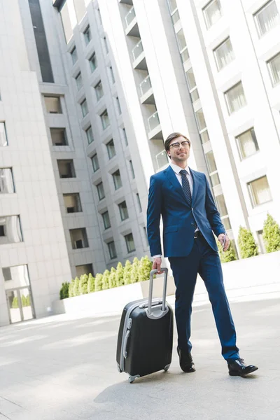 Handsome young businessman in stylish suit with luggage walking by business district — Stock Photo