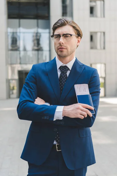 Thoughtful young businessman with crossed arms holding flight tickets — Stock Photo