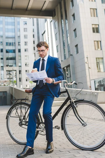 Businessman in stylish suit with newspaper and paper cup of coffee leaning on bicycle — Stock Photo