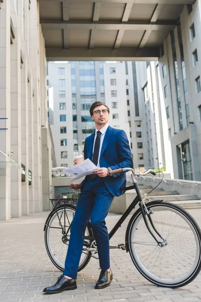 Jovem empresário em terno elegante com jornal e xícara de papel de café encostado na bicicleta — Fotografia de Stock