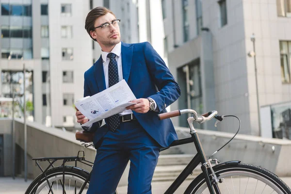 Young businessman in stylish suit with newspaper leaning on bicycle — Stock Photo