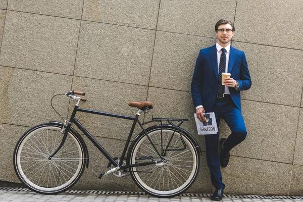 Handsome young businessman in stylish suit with newspaper and bicycle leaning on wall — Stock Photo