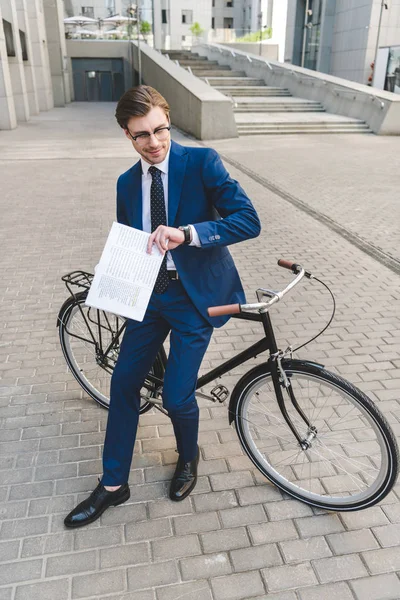 Handsome young businessman in stylish suit with newspaper leaning on bicycle — Stock Photo