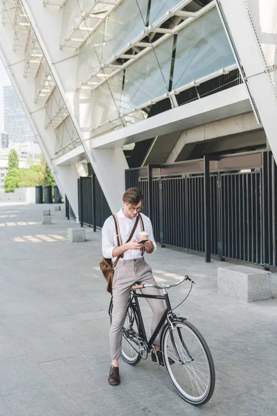 Joven guapo con café para ir usando el teléfono inteligente mientras está sentado en bicicleta - foto de stock