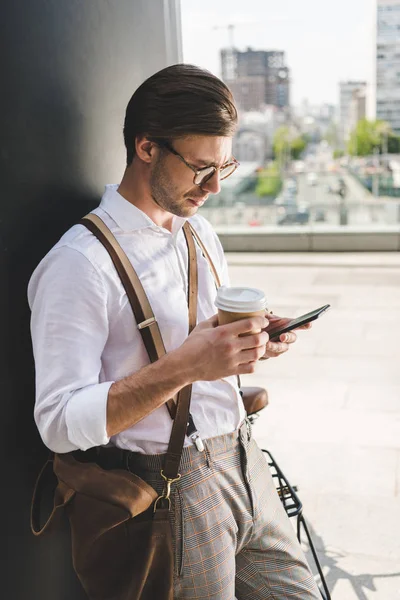 Стильный молодой человек с бумажной чашкой кофе будет ходить со смартфоном — стоковое фото