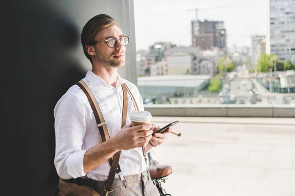 Jeune homme réfléchi avec du café pour aller à l'aide d'un smartphone et détourner les yeux — Photo de stock