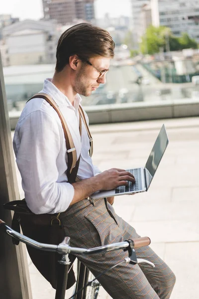 Side view of handsome young man working with laptop while leaning on vintage bicycle — Stock Photo