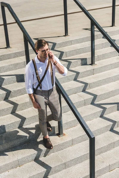 High angle view of stylish young man with folded laptop walking on stairs on city street and talking by phone — Stock Photo
