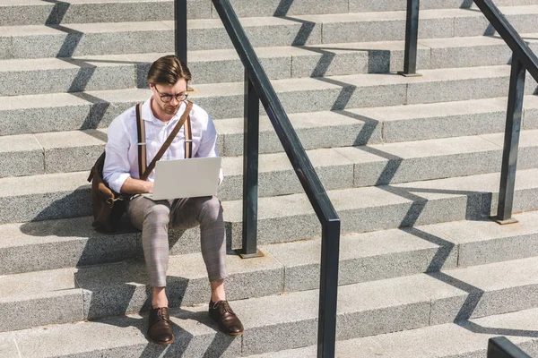 Handsome young man working with laptop on stairs on city street — Stock Photo