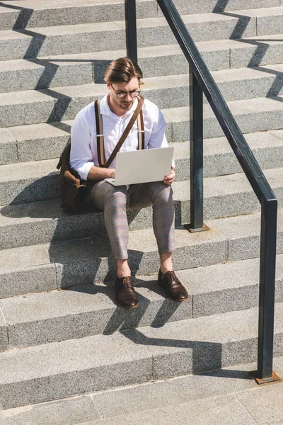 Attractive young man working with laptop on stairs on city street — Stock Photo