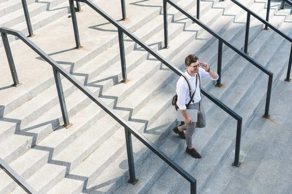 Vue grand angle de jeune homme élégant marchant sur les escaliers avec ordinateur portable sur la rue de la ville — Photo de stock