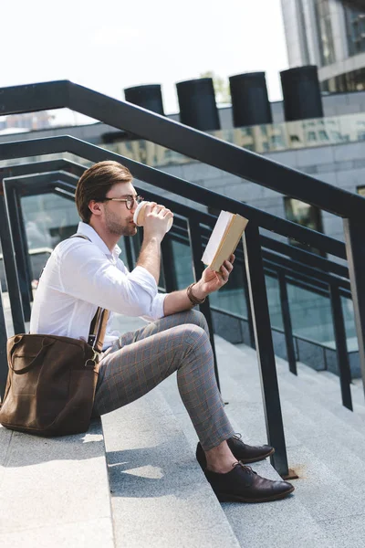 Красивый молодой человек пьет кофе из бумажной чашки и читает книгу на лестнице на городской улице — стоковое фото