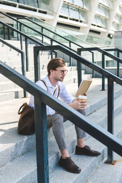 Jovem concentrado com xícara de papel de livro de leitura de café em escadas na rua da cidade — Fotografia de Stock