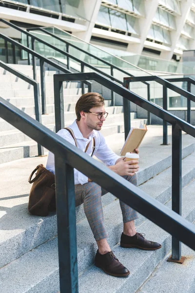 Joven concentrado con taza de papel de libro de lectura de café en las escaleras en la calle de la ciudad - foto de stock