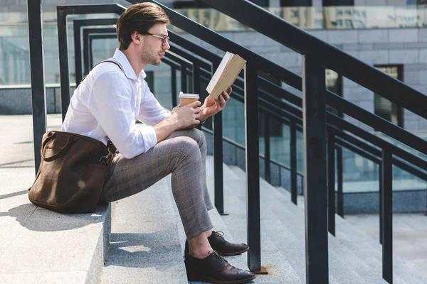 Vista lateral del joven guapo con taza de papel de libro de lectura de café en las escaleras en la calle de la ciudad - foto de stock
