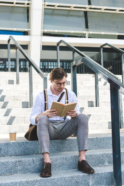 Ориентированный молодой человек с бумажной чашкой кофе чтение книги на лестнице на городской улице — стоковое фото
