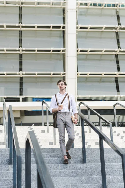 Joven con estilo bajando escaleras frente al estadio con libro y café para llevar - foto de stock