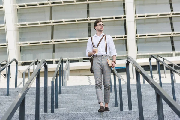 Atractivo joven bajando escaleras frente al estadio con libro y café para llevar - foto de stock