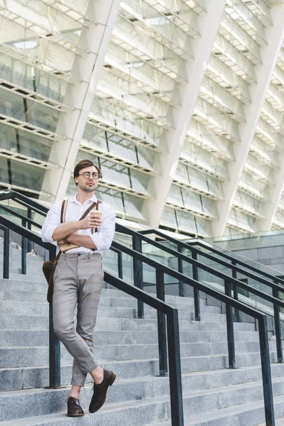 Стильный молодой человек, стоящий на лестнице перед стадионом с книгой и кофе, чтобы пойти — стоковое фото