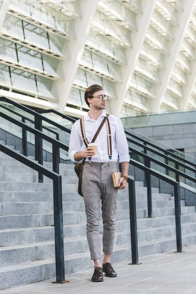 Jovem atraente andando na frente do estádio com livro e café para ir — Fotografia de Stock