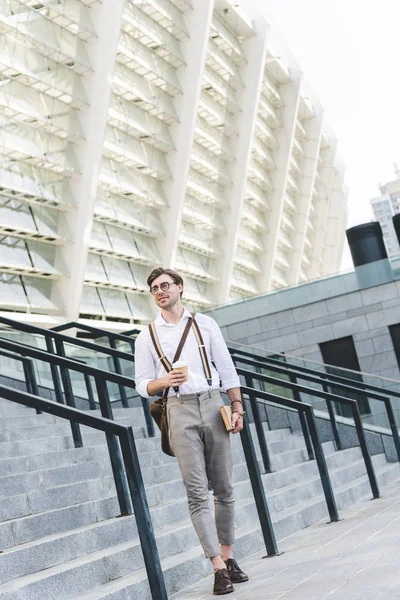 Стильный молодой человек, идущий перед стадионом с книгой и кофе, чтобы пойти — стоковое фото