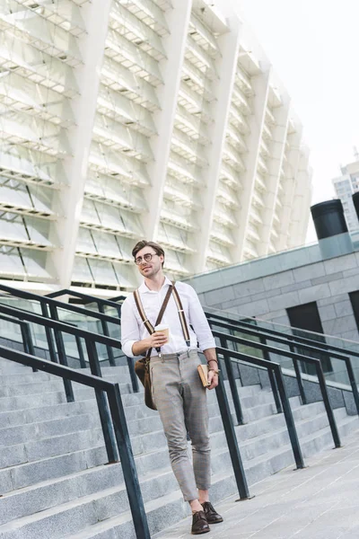 Joven con estilo caminando frente al estadio con libro y café para llevar - foto de stock