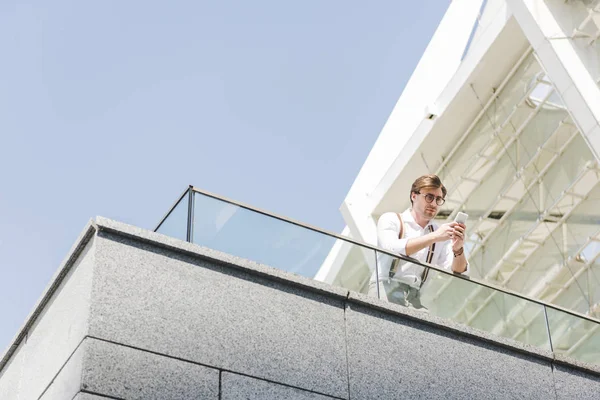 Вид снизу на привлекательного молодого человека с помощью смартфона на балконе — стоковое фото