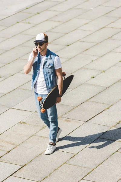 Vista de ángulo alto de joven patinador guapo caminando con longboard en la mano y hablando por teléfono - foto de stock