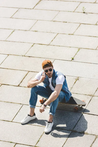 Vista de ángulo alto del joven patinador guapo sentado en longboard y hablando por teléfono - foto de stock