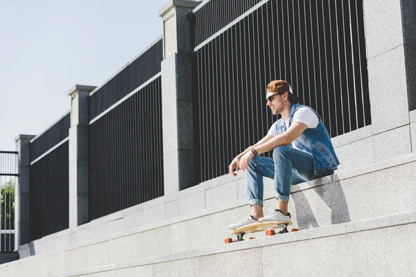 Стильный молодой человек в джинсовой одежде сидит на лестнице с длинной доской, глядя в сторону — стоковое фото