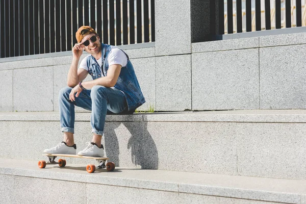 Sorrindo jovem patinador sentado em escadas com longboard olhando para longe — Fotografia de Stock