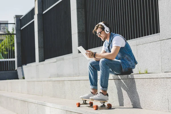 Stylish young man with skateboard listening music with headphones and tablet on street — Stock Photo