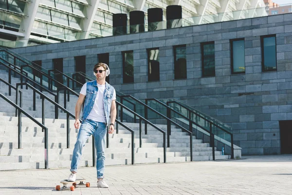 Handsome young man in headphones with skateboard in front of stadium — Stock Photo