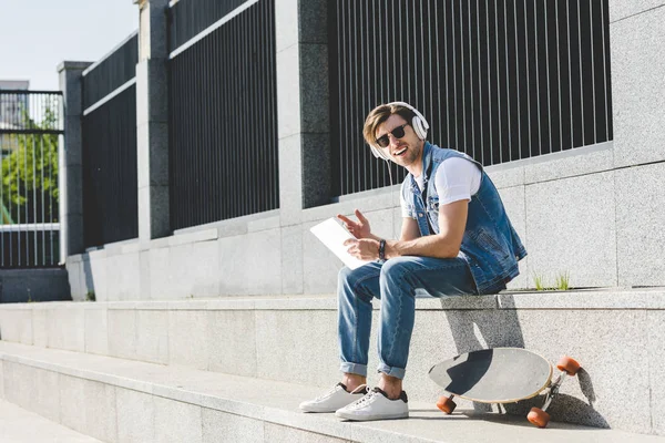 Jovem feliz com skate ouvindo música com fones de ouvido e tablet na rua — Fotografia de Stock