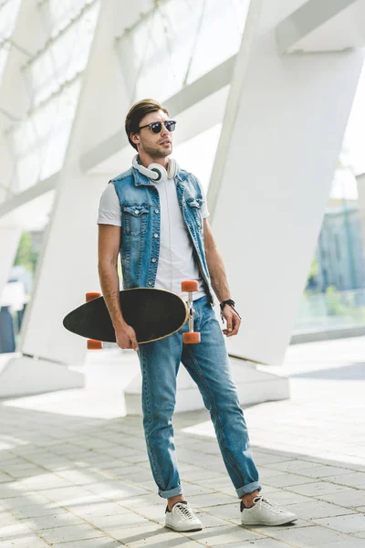 Jovem skatista em roupas jeans com longboard e fones de ouvido — Fotografia de Stock
