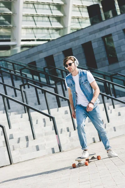 Handsome young skater in headphones riding longboard in front of stadium — Stock Photo