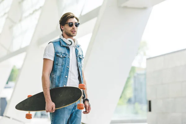 Stylish young skater in denim clothes with longboard and headphones — Stock Photo