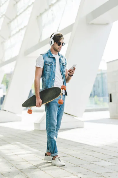 Stylish young man with skateboard using smartphone on street of modern city — Stock Photo