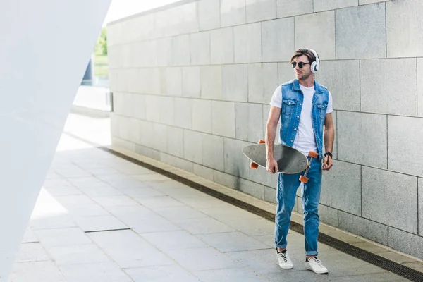 Lächelnder junger Mann mit Kopfhörern und Skateboard läuft durch die Straße — Stockfoto