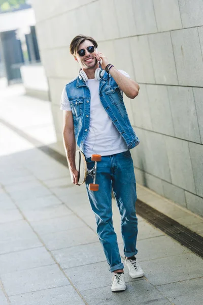 Smiling young man with skateboard talking by phone — Stock Photo