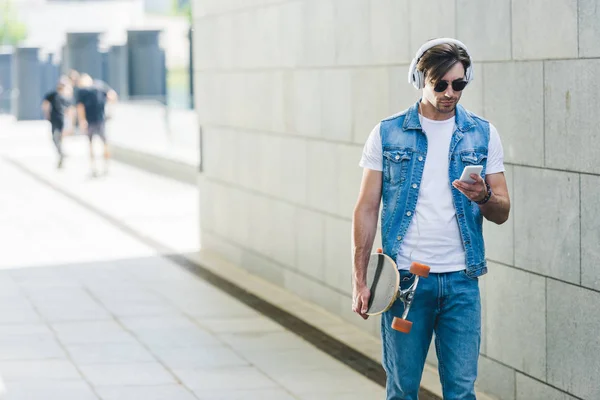 Jovem bonito em fones de ouvido com skate e smartphone — Fotografia de Stock