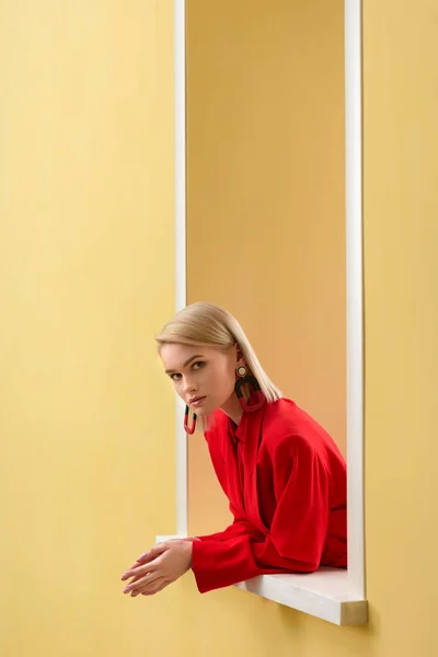 Vista laterale della donna bionda elegante in abito rosso guardando fuori finestra decorativa — Foto stock