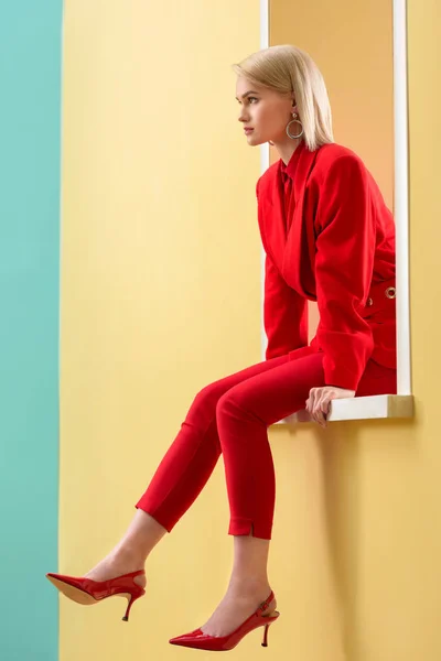 Side view of beautiful blond woman in stylish red suit and shoes sitting on decorative window — Stock Photo