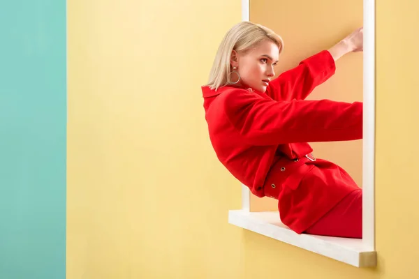 Side view of beautiful blond woman in stylish red suit sitting on decorative window — Stock Photo