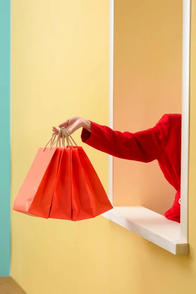 Partial view of stylish woman in red clothing with red shopping bags outstretching hand out decorative window — Stock Photo