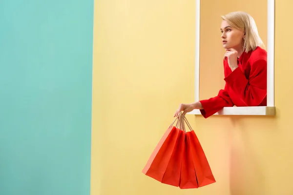 Mulher elegante pensativo em roupas vermelhas com sacos de compras vermelhos olhando para fora janela decorativa — Fotografia de Stock