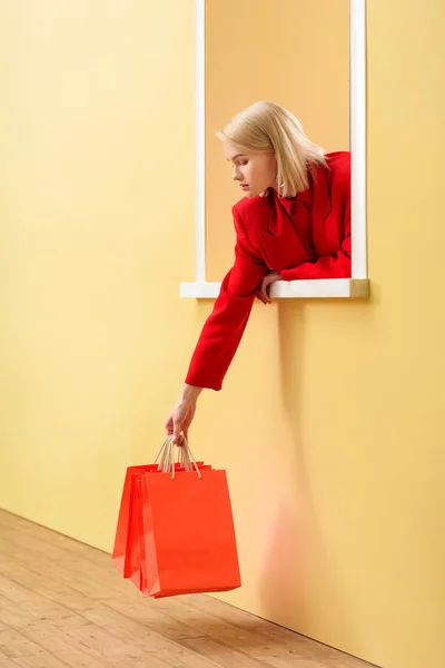 Jeune femme à la mode en vêtements rouges avec des sacs à provisions rouges regardant par la fenêtre décorative — Photo de stock